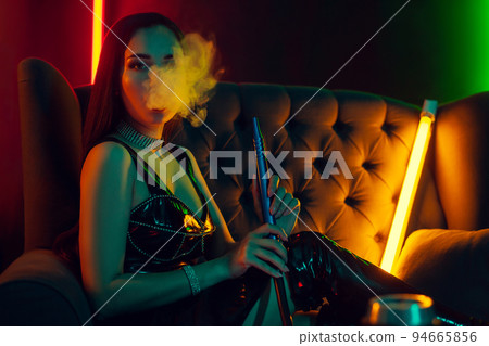 Sexy brunette model is smoking a hookah exhaling a smoke at a luxury night club. Sexy brunette model is smoking a hookah exhaling a smoke at a luxury night club. 94665856
