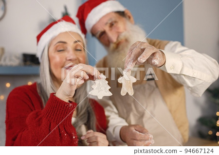 Senior man and his wife baking cookies together. Old bearded man standing together near his wife with grey hair. Man wearing white sweater and woman red one. 94667124