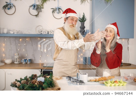 Senior man and his wife cooking together. Old bearded man standing together near his wife with grey hair. Man wearing white sweater and woman red one. 94667144