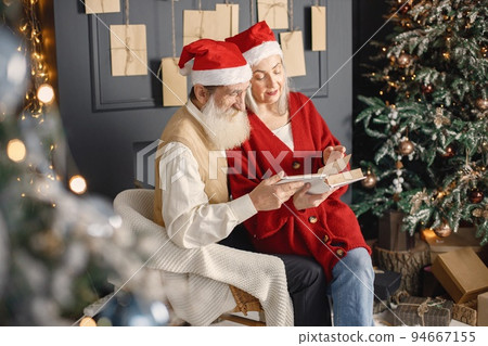 Senior man celebrating christmas with his wife near christmas tree. Old bearded man with his wife with grey hair sitting and reading a book. Man wearing white sweater and woman red one, have a red Senior man celebrating christmas with his wife near christmas tree. Old bearded man with his wife with grey hair sitting and reading a book. Man wearing white sweater and woman red one, have a red 94667155