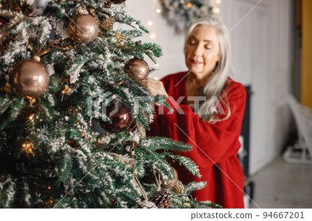 Charming old female is preparing for New Year. Senior woman standing near christmas tree. Woman has gray hair and wearing red sweater. 94667201