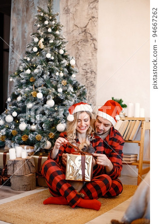 Romantic couple with their french bulldog celebrating Christmas on a floor near Christmas tree. Blonde woman and brunette man wearing plaid pajamas. Wife and husband opening present box. Romantic couple with their french bulldog celebrating Christmas on a floor near Christmas tree. Blonde woman and brunette man wearing plaid pajamas. Wife and husband opening present box. 94667262