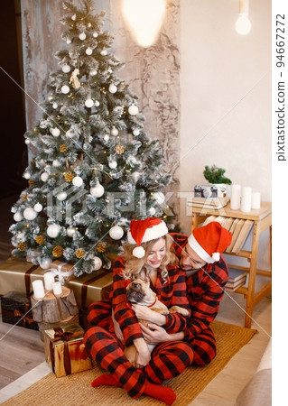 Romantic couple celebrating Christmas on a floor near Christmas tree. Blonde woman and brunette man wearing plaid pajamas. Wife and husband hugging their french bulldog. Romantic couple celebrating Christmas on a floor near Christmas tree. Blonde woman and brunette man wearing plaid pajamas. Wife and husband hugging their french bulldog. 94667272