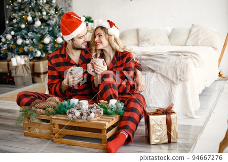 Romantic couple celebrating Christmas on a floor near Christmas tree. Blonde woman and brunette man wearing plaid pajamas. Romantic couple drinking a tea from a mugs. 94667276