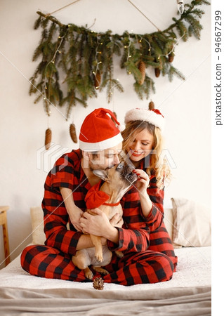 Lovely couple celebrating christmas with dog on a bed. Blonde woman and brunette man wearing plaid pajamas. French bulldog with his family. 94667299
