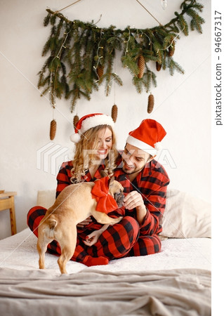Lovely couple celebrating christmas with dog on a bed. Blonde woman and brunette man wearing plaid pajamas. French bulldog with his family. 94667317
