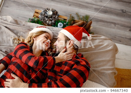 Lovely couple celebrating christmas on a bed. Blonde woman and brunette man wearing plaid pajamas. Romantic couple hugging. Lovely couple celebrating christmas on a bed. Blonde woman and brunette man wearing plaid pajamas. Romantic couple hugging. 94667340