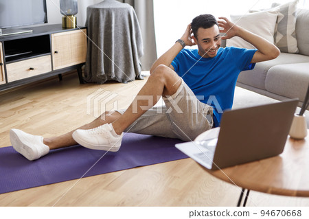 Young arab guy exercising at home, having online fitness video and working on abs strength, looking at laptop screen Young arab guy exercising at home, having online fitness video and working on abs strength, looking at laptop screen 94670668