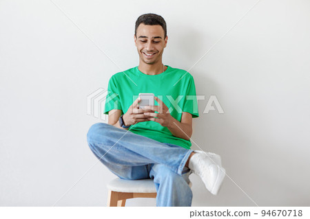 People and technology. Positive arab guy using smartphone sitting on chair over light studio wall background 94670718