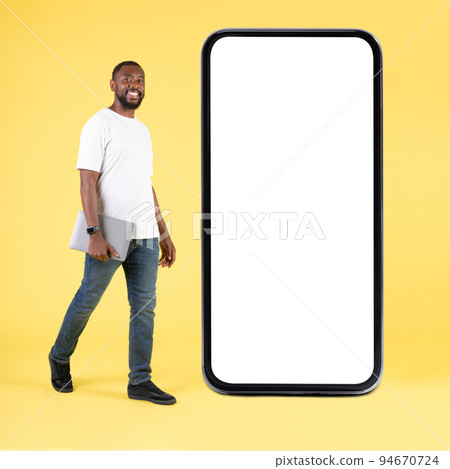 Happy Black Guy Near Large Smartphone Holding Laptop, Yellow Background Happy Black Guy Near Large Smartphone Holding Laptop, Yellow Background 94670724