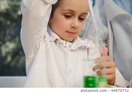 Beautiful mischievous child girl holding a glass pipette, and dripping few drops of green chemical solution from a flask into a test tube during a chemistry lesson, in the school science laboratory 94670752