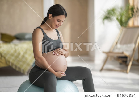 Smiling Beautiful Pregnant Woman Sitting On Fitball At Home And Embracing Belly 94670854