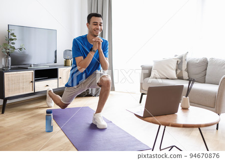 Happy arab man doing forward lunge exercises while watching online workout tutorial via laptop, training at home 94670876