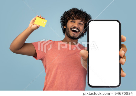 Happy indian guy showing cell phone and credit card, mockup 94670884
