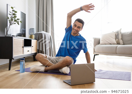 Positive arab man having fitness class online at home, using laptop, watching sport videos on Internet and stretching Positive arab man having fitness class online at home, using laptop, watching sport videos on Internet and stretching 94671025