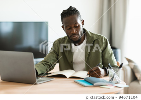 Focused african american man sitting at desk working on laptop and taking notes in notebook, studying online at home 94671104