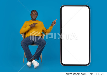 Positive black man sitting on chair by huge smartphone, mockup 94671176