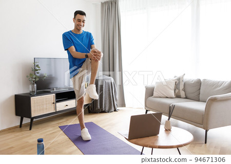Happy arab man stretching his leg in front of laptop during domestic training, following online video tutorial Happy arab man stretching his leg in front of laptop during domestic training, following online video tutorial 94671306