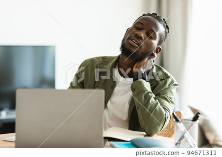 Tired black man suffering from neck spasm while working on laptop and rubbing his muscles, sitting at desk Tired black man suffering from neck spasm while working on laptop and rubbing his muscles, sitting at desk 94671311