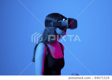 Confident brunette young woman in virtual reality headset pointing in the air while standing in a studio space. Using VR technologies. Confident brunette young woman in virtual reality headset pointing in the air while standing in a studio space. Using VR technologies. 94671529