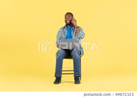African American Guy Using Laptop Talking On Cellphone, Yellow Background 94672323