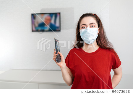 A young woman in a medical mask turns on the remote control TV, which shows false news. Copy space. The concept of disinformation and fictional news A young woman in a medical mask turns on the remote control TV, which shows false news. Copy space. The concept of disinformation and fictional news 94673638