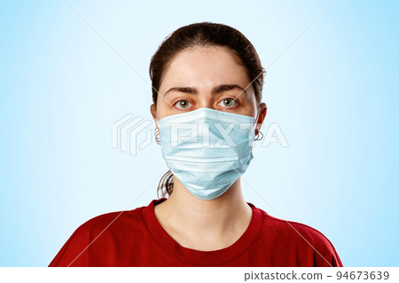 Portrait of a brunette woman in a medical mask and a red t-shirt, on a light blue background. Copy space. Concept of pandemic and protection against viral infection Portrait of a brunette woman in a medical mask and a red t-shirt, on a light blue background. Copy space. Concept of pandemic and protection against viral infection 94673639