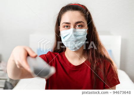 Portrait of a Young woman in a medical mask holding a TV remote control. Close up. The concept of disinformation and false news Portrait of a Young woman in a medical mask holding a TV remote control. Close up. The concept of disinformation and false news 94673648