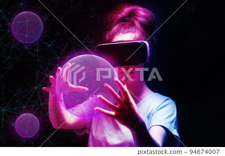 Portrait of young woman in VR glasses creates 3D simulation of pink mesh sphere. Dark background with neon abstracts. The concept of virtual reality and metaverse 94674007