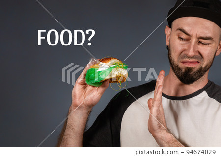 The concept of fast food and ecology. A man in a cap and a beard, holding a hamburger with a plastic filling, and with disgust refuses it. Text FOOD. lack background The concept of fast food and ecology. A man in a cap and a beard, holding a hamburger with a plastic filling, and with disgust refuses it. Text FOOD. lack background 94674029