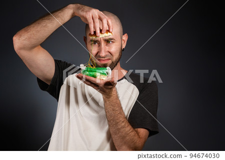 The concept of fast food and ecology. A bald man with a beard looks at the stuffing of a disassembled hamburger with horror. Dark background. Copy space The concept of fast food and ecology. A bald man with a beard looks at the stuffing of a disassembled hamburger with horror. Dark background. Copy space 94674030