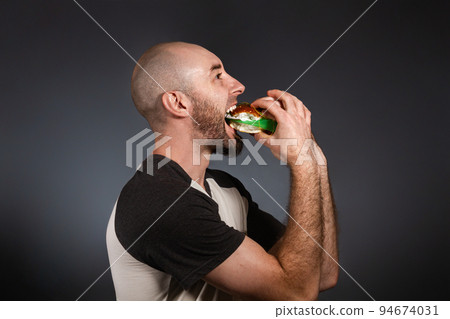 The concept of fast food and ecology. A bald man with a beard, happily eating a hamburger stuffed with garbage. Copy space. Dark background The concept of fast food and ecology. A bald man with a beard, happily eating a hamburger stuffed with garbage. Copy space. Dark background 94674031