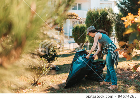 The concept of environmental pollution and Earth day. Woman volunteer doing cleaning in the Park in front of the house. She's putting the garbage bags in one pile. Close up The concept of environmental pollution and Earth day. Woman volunteer doing cleaning in the Park in front of the house. She's putting the garbage bags in one pile. Close up 94674263