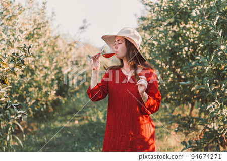 Wine tasting and relaxing in the garden. Young beautiful brunette in a red dress and hat drinking a glass of wine. There are fruit trees in the background Wine tasting and relaxing in the garden. Young beautiful brunette in a red dress and hat drinking a glass of wine. There are fruit trees in the background 94674271