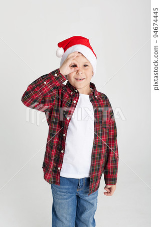 A boy in a red T-shirt and a Santa Claus hat is smiling. Funny kid in the studio for Christmas on a white background. The European kid smiles and makes a face. New Year 94674445