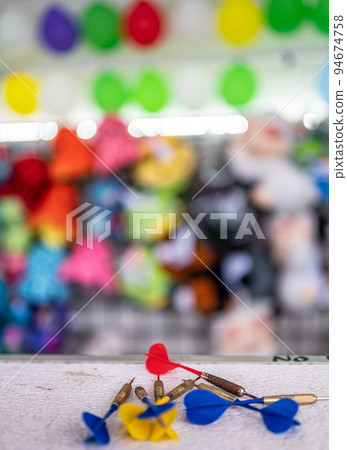 selective focus on darts sitting on a ledge with a wall of balloon targets and prizes that a player can win at a fair. selective focus on darts sitting on a ledge with a wall of balloon targets and prizes that a player can win at a fair. 94674758