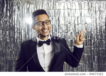 Smiling elegant young man in black tuxedo pointing sideways with index finger on silver background. 94675361