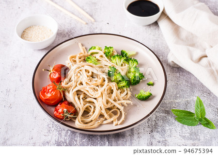 Fried udon noodles in soy sauce with tomatoes, broccoli and sesame seeds on a plate on the table 94675394