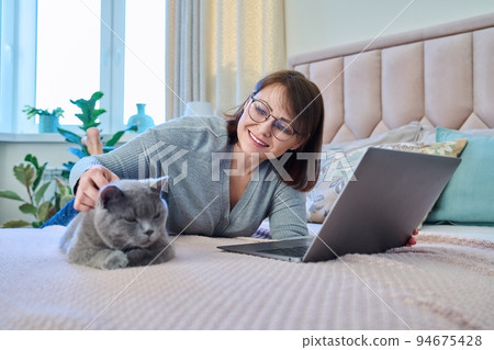 Woman together with cat lying on bed at home, female using laptop Woman together with cat lying on bed at home, female using laptop 94675428