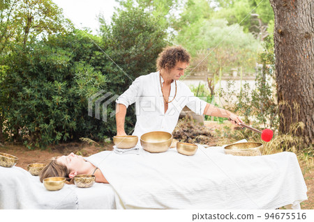 A man wearing in white, master of healing sound massage, doing sound therapy with tibetan singing bowl to a young woman A man wearing in white, master of healing sound massage, doing sound therapy with tibetan singing bowl to a young woman 94675516