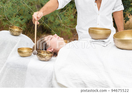 Young woman laying on a massage bed with tibetan singing bowl outdoor, male hand holding a felt clapper 94675522