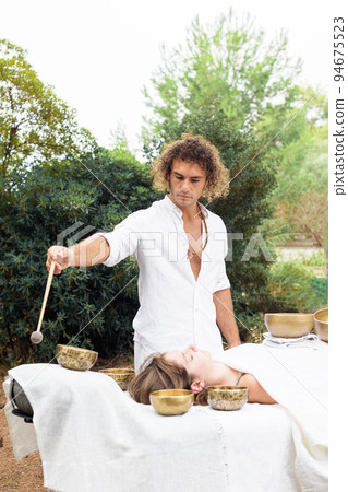 A man wearing in white, master of healing sound massage, doing sound therapy with tibetan singing bowl to a young woman 94675523