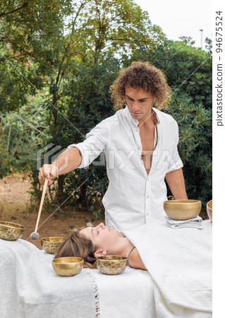 A man wearing in white, master of healing sound massage, doing sound therapy with tibetan singing bowl to a young woman 94675524