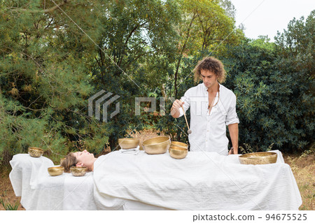 A man wearing in white, master of healing sound massage, doing sound therapy with tibetan singing bowl to a young woman 94675525