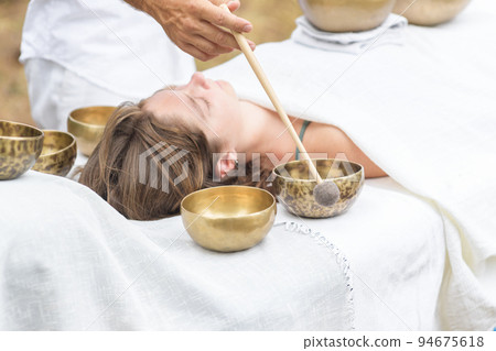 Young woman laying on a massage bed with tibetan singing bowl outdoor, male hand holding a felt clapper 94675618