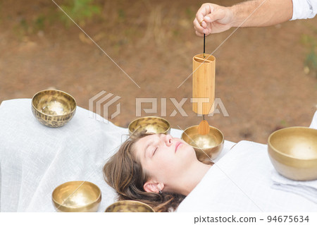 Male hand holding a bamboo Koshi chime during sound healing therapy with tibetan singing bowls over young woman outdoor 94675634