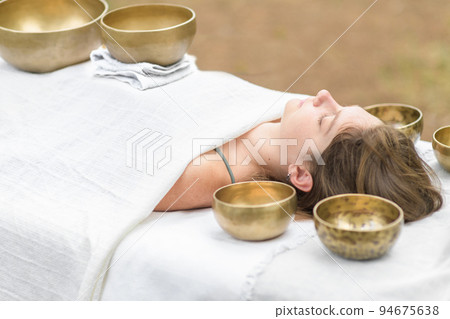 Young woman laying on a massage bed with tibetan singing bowl outdoor. Nepal copper singing bowl sound massage 94675638