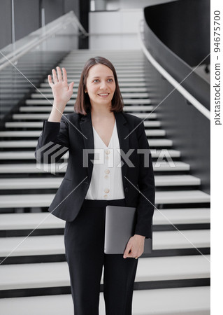 business woman in a formal suit with a laptop in the office of the business center stands on the steps and greets colleagues 94675700