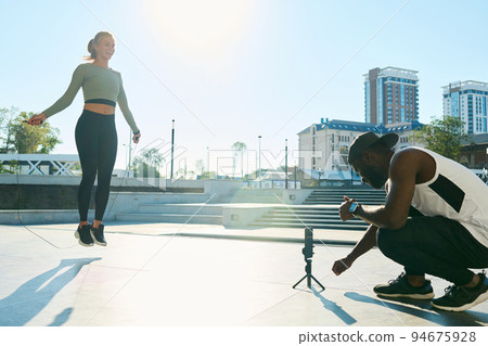 Happy young woman jumping over skipping rope while man shooting video Happy young woman jumping over skipping rope while man shooting video 94675928