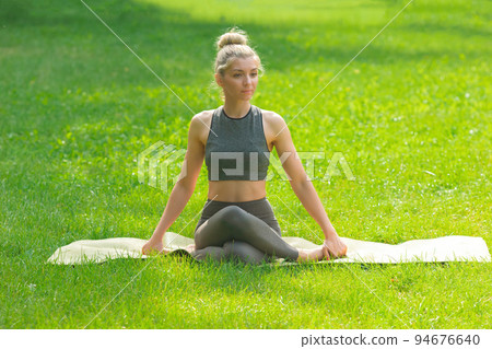 A slender woman sitting on the green grass in the park, performing body twists A slender woman sitting on the green grass in the park, performing body twists 94676640